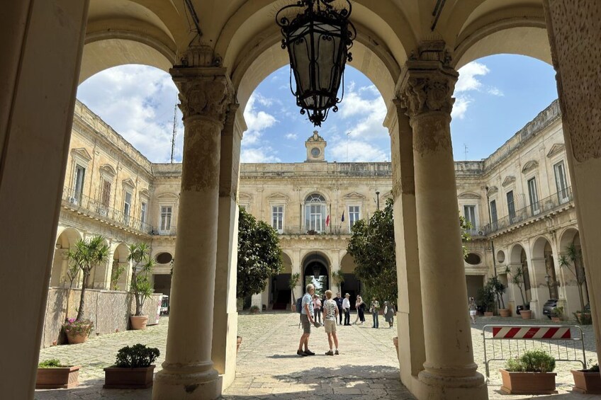 Cortile aperto con archi, un edificio centrale con una torre dell'orologio e persone che passeggiano. 1