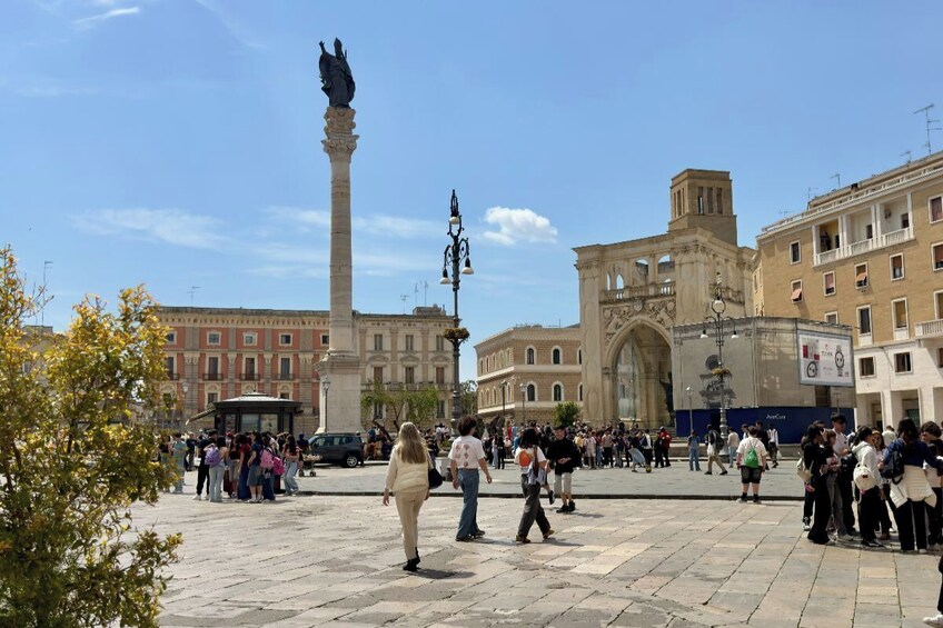 Una piazza pubblica con un monumento imponente, edifici storici e persone che passeggiano. 2