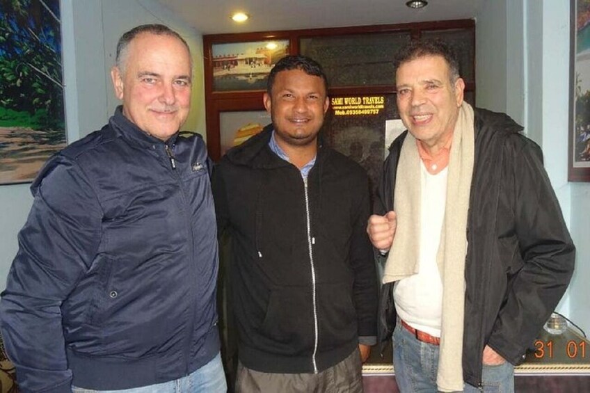 Three men standing indoors with a sign reading 'SAM WORLD TRAVELS' in the background. 4