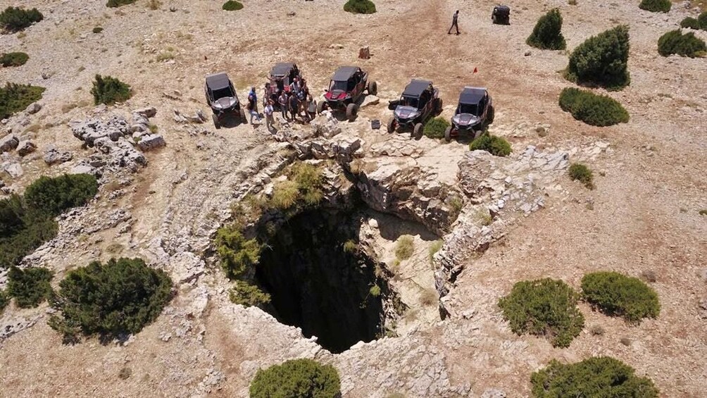 A group of off-road vehicles parked around a large sinkhole in a rocky terrain. 1