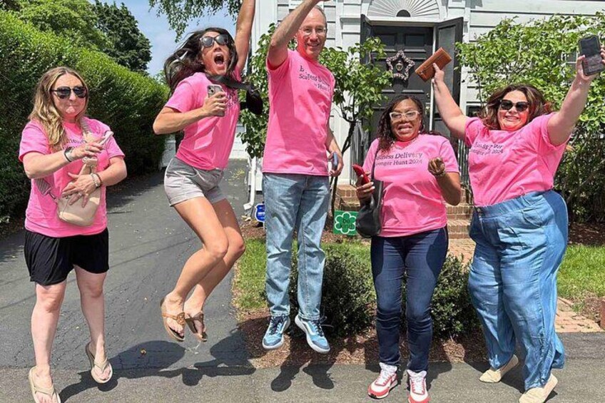 A group of people in pink shirts, some holding phones, in front of a building with a white facade and a black door. 3
