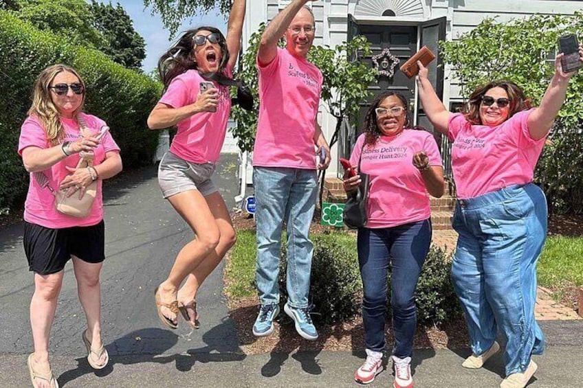 Un grupo de personas con camisetas rosas, algunas sosteniendo teléfonos, de pie frente a un edificio con jardín. 3