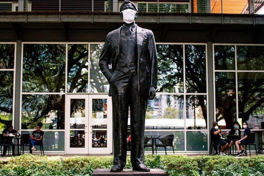Una estatua de un hombre con traje y sombrero, sobre un pedestal en un parque con árboles y un edificio al fondo. 4