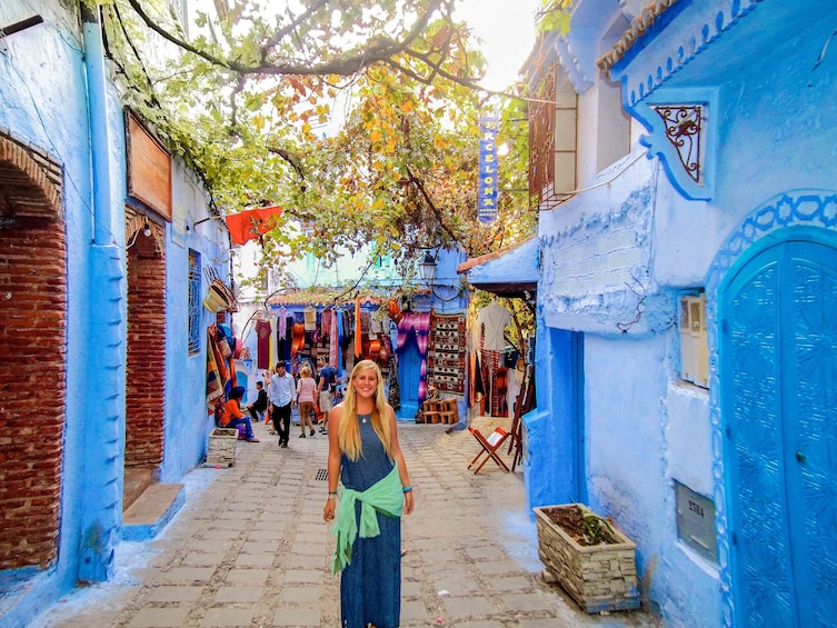 A narrow street with blue buildings, a woman in a blue dress, and hanging fabrics. 1