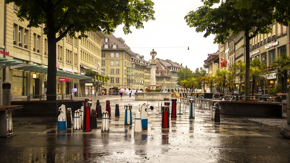A wet city square with a fountain, trees, and buildings. 4