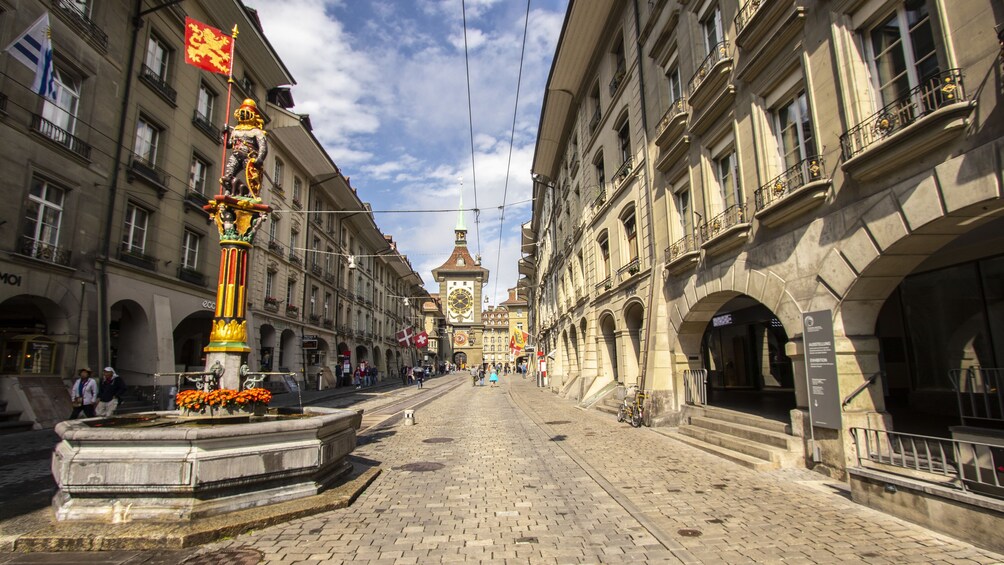 A cobblestone street lined with historic buildings, a fountain, and a clock tower in the background. 3