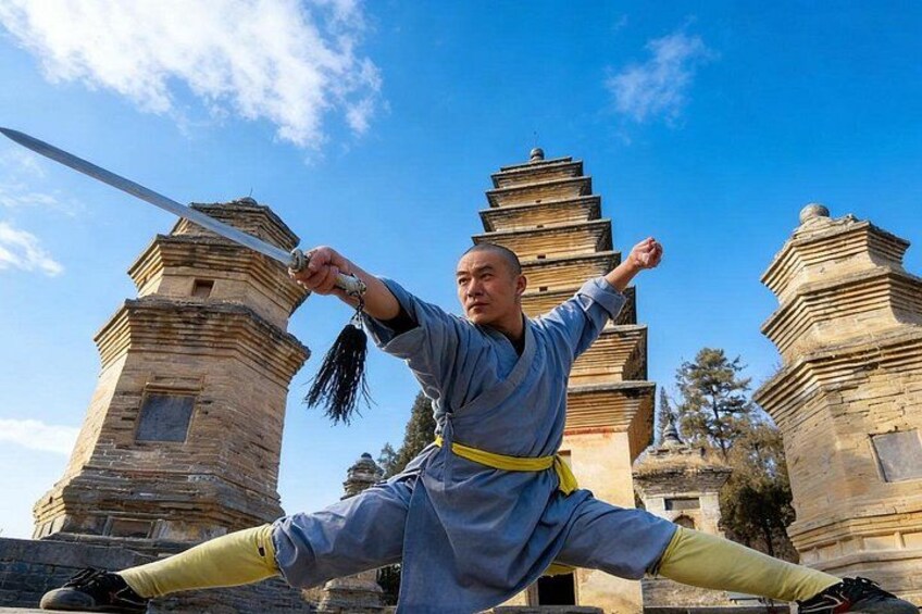 A person in traditional martial arts attire performing a split with a sword in front of ancient stone pagodas. 1