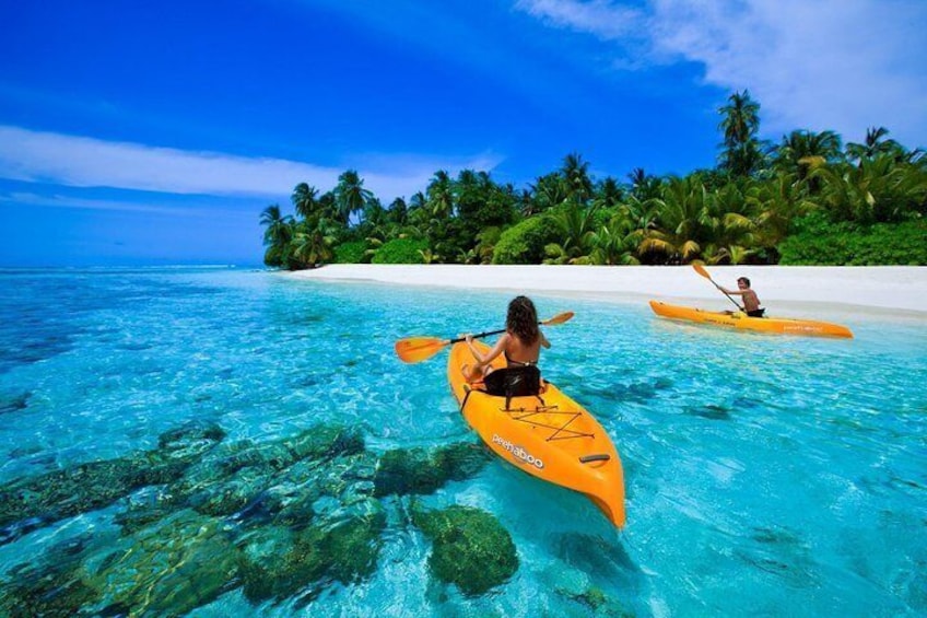 Two people kayaking on clear turquoise water with a white sandy beach and palm trees in the background. 1