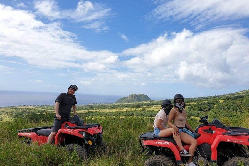 Two people on ATVs in a grassy field with a mountainous landscape in the background. 3