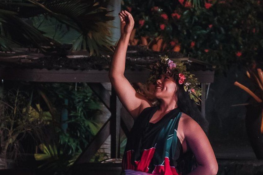 A person with a flower crown, wearing a sleeveless dress, in an outdoor setting with plants and a wooden structure. 2