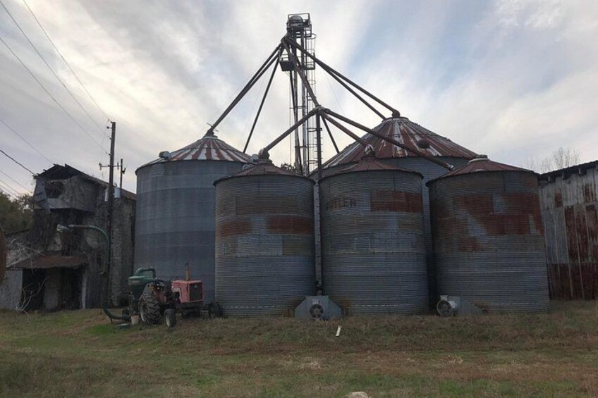 Tre grandi silos metallici con una torre centrale, immersi in un'area erbosa con un edificio fatiscente e un trattore. 2