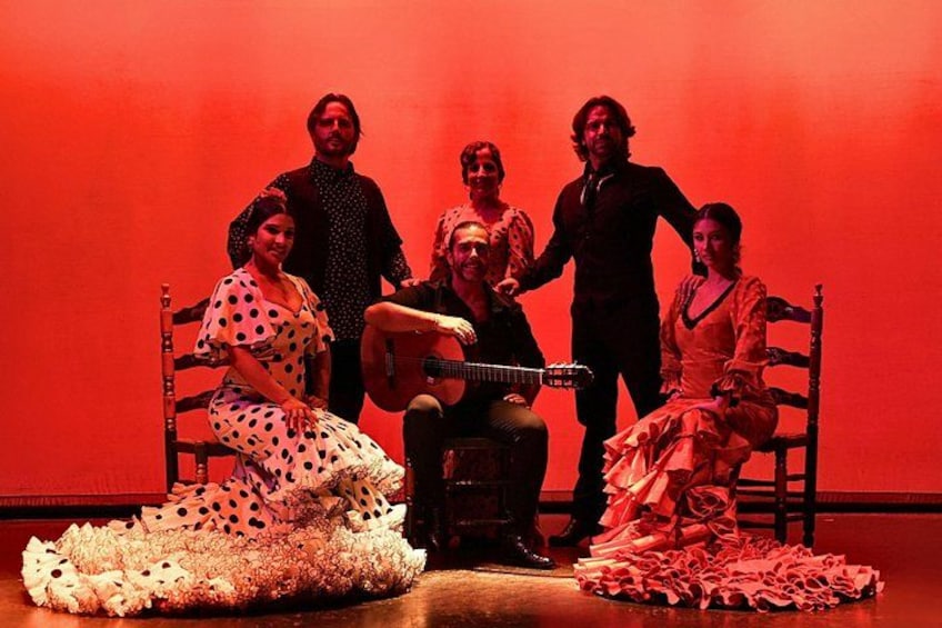 Un grupo de personas con vestuario de época en un escenario, con una guitarra, iluminación roja y arreglos florales. 3