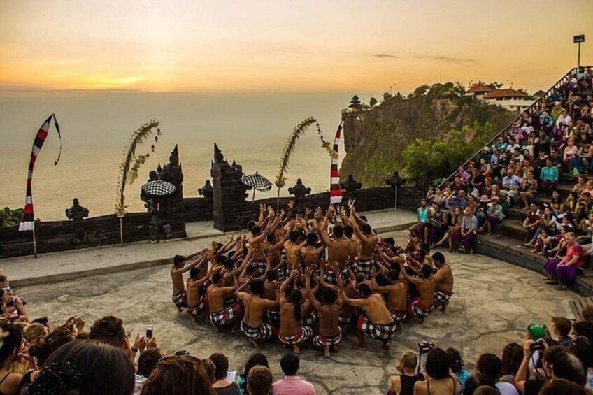 A traditional dance performance by a group of men in a circular formation, with an audience seated on steps and a coastal backdrop. 4