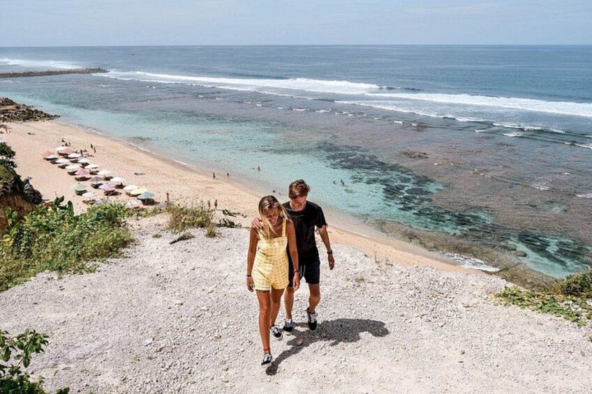 A beach with a rocky path leading to the shore, a sandy beach with umbrellas, and the ocean in the background. 2