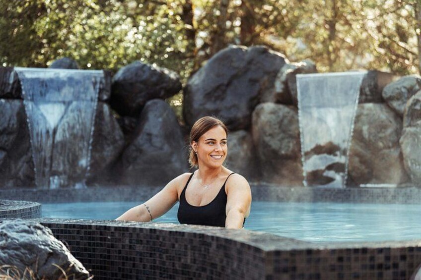 A person in a black swimsuit at a hot spring with a waterfall and rocks in the background. 5