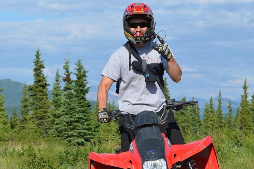 A person on a red ATV in a grassy field with trees and mountains in the background. 5