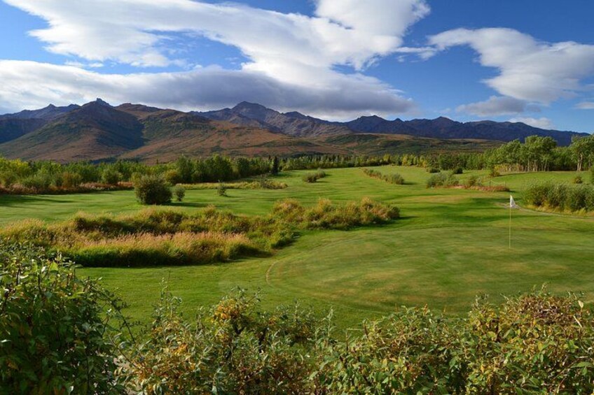 En golfbane med et flag, grønne bakker og bjerge i baggrunden. 1
