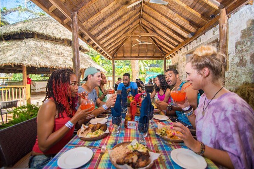 A group of people dining at an outdoor restaurant with a thatched roof and colorful tablecloths. 2