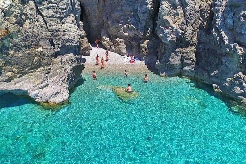 Una playa rocosa con agua turquesa cristalina, gente nadando y una pequeña isla en el centro. 2
