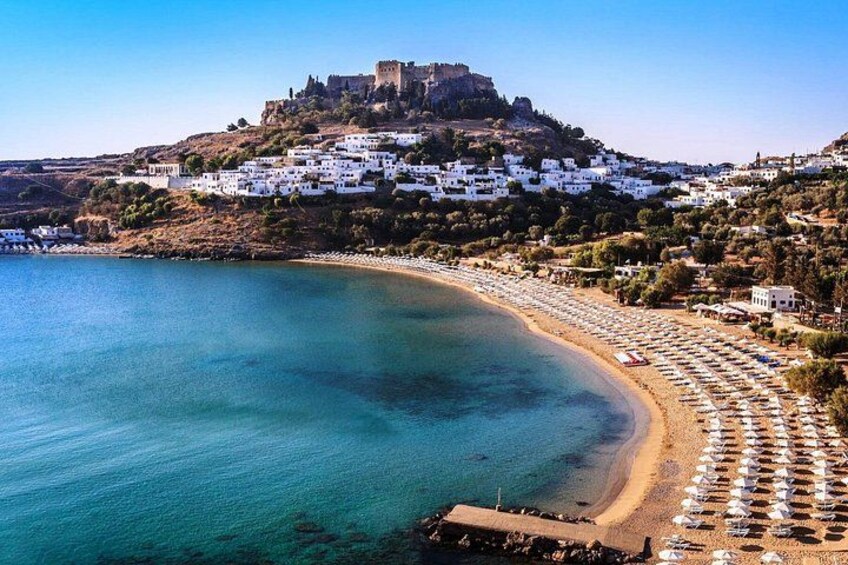A coastal town with a castle on a hill, a beach with umbrellas, and clear blue waters. 1