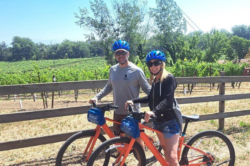 Dos personas en bicicleta con casco, un viñedo al fondo y una valla de madera. 4