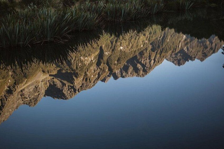 A calm body of water reflecting a rocky shoreline with sparse vegetation. 2