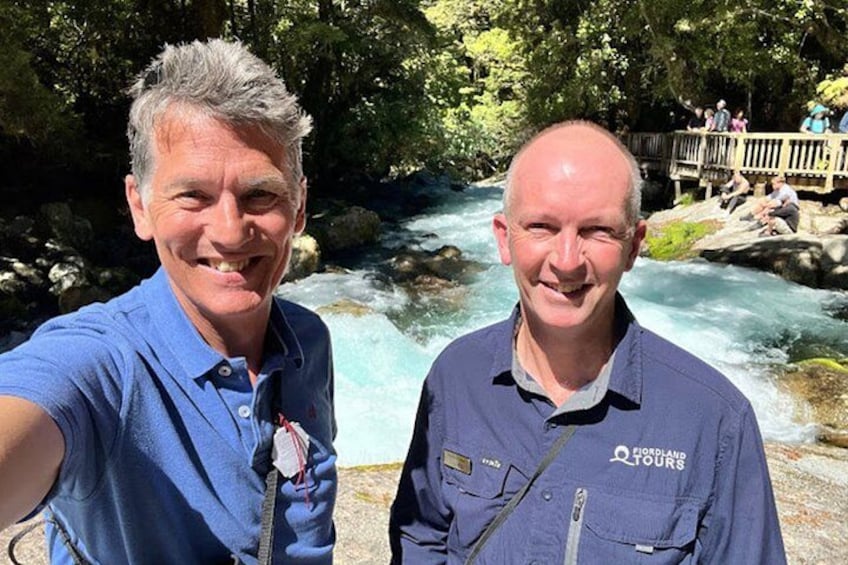 Two people standing by a flowing river with a wooden bridge in the background. 5