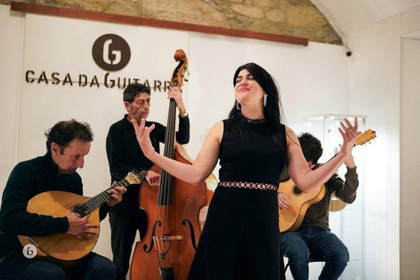 Une personne chante accompagnée d’une guitare, d’une contrebasse et d’une autre guitare, dans une pièce où est affiché un panneau indiquant « GASA DA GUITARRA ». 2