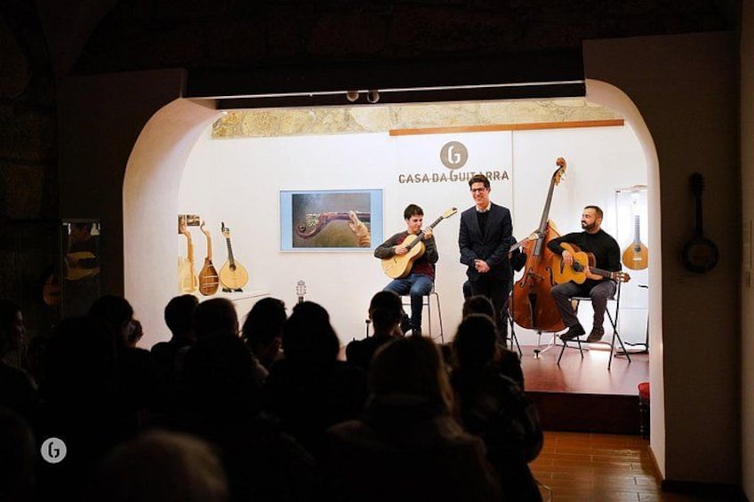 Et band optræder på scenen med guitarer og en cello, i et lokale med en hvid væg og et indrammet billede. 2