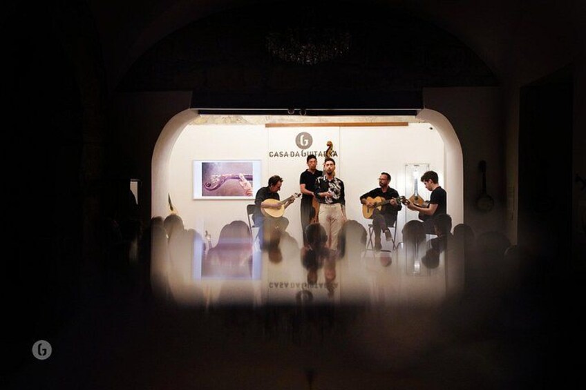 Un escenario con un grupo de personas, algunas tocando guitarras, en una habitación poco iluminada con un fondo blanco y un logotipo. 3