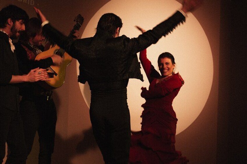 Un grupo de personas, una tocando la guitarra, en una habitación tenuemente iluminada con una gran fuente de luz circular al fondo. 2