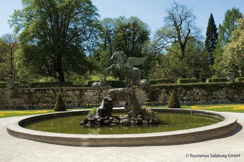 A fountain with a statue of a horse and rider, surrounded by a circular stone border, set in a park with trees and a stone wall in the background. 3