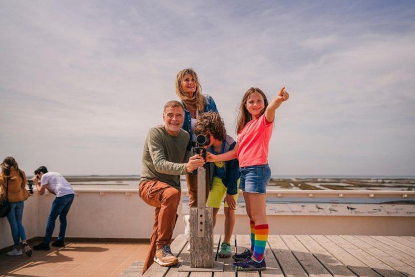 A family on a wooden deck by the sea, with a photographer and a child pointing. 1
