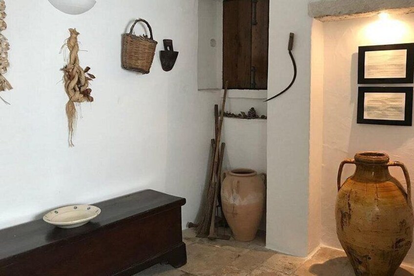 A rustic interior with a large clay pot, a wooden bench, and wall-mounted baskets. 1