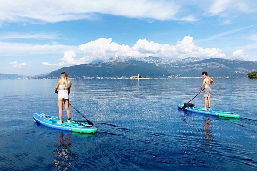 To personer står på paddleboards på en rolig sø med bjerge i baggrunden. 4