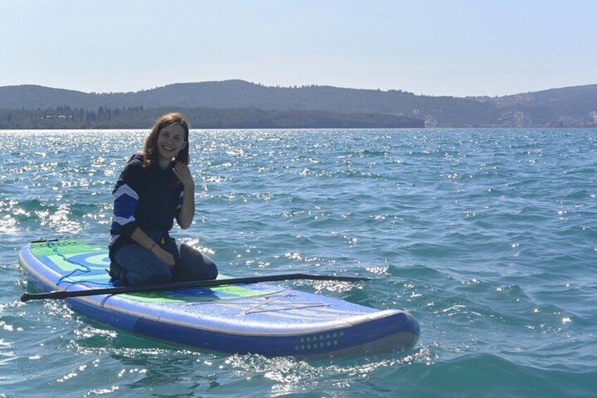 En person sidder på et blåt paddleboard i stille vand med et bakket landskab i baggrunden. 2