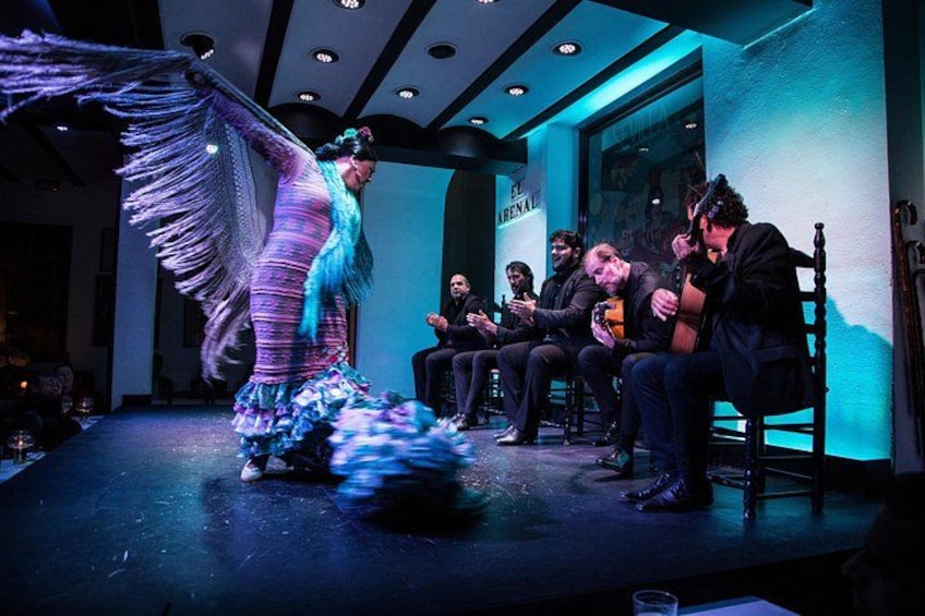 A performer in a colorful dress with a wing-like extension on stage, seated audience, and a building with a sign. 2