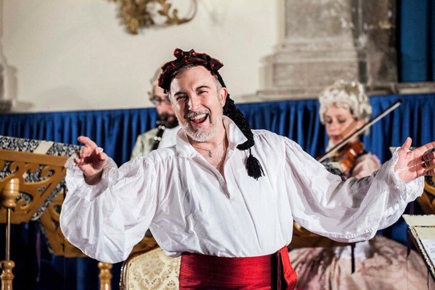 Une personne vêtue d’une chemise blanche et d’une écharpe rouge, peut-être un chef d’orchestre, avec un violoniste en arrière-plan. 2