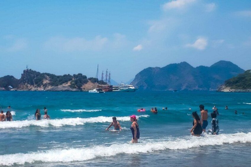 A beach with people swimming and standing in the water, boats in the background, and mountains in the distance. 2