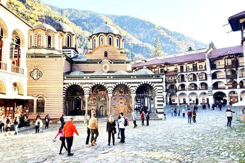 A historic square with a church, people walking, and a mountainous backdrop. 1