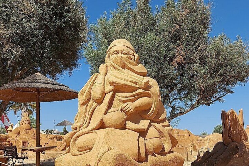 A large sand sculpture of a seated figure with a headscarf, surrounded by trees and a thatched umbrella. 2