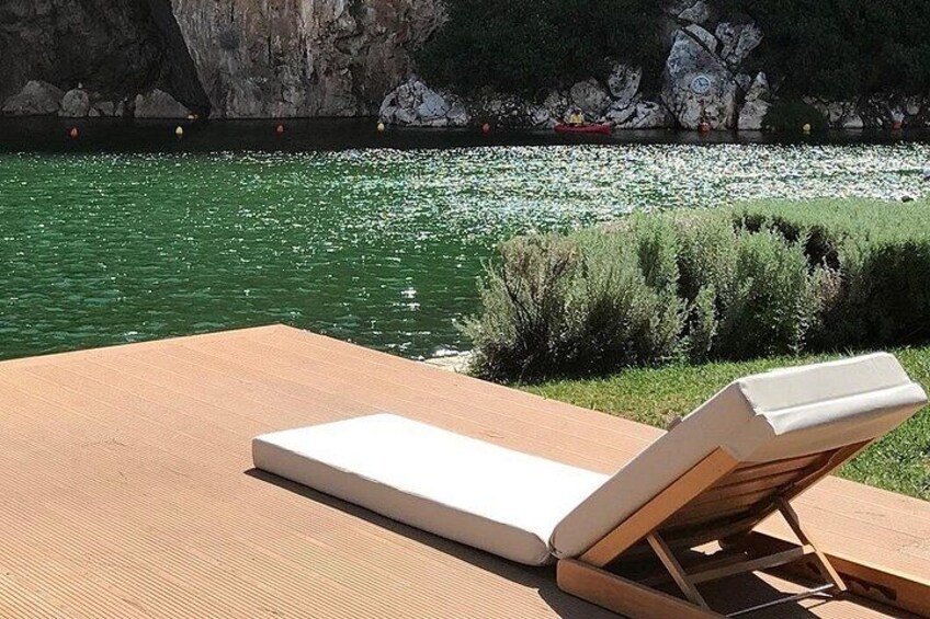 A lounge chair on a wooden deck by a calm body of water with rocks and greenery in the background. 4