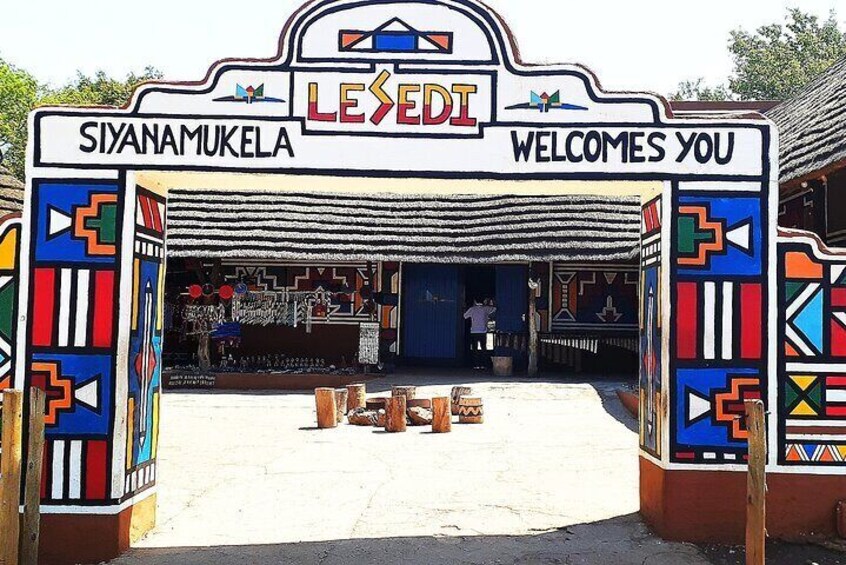 A colorful entrance archway with 'Siyananukela' and 'Lesedi' written on it, leading to a building with a thatched roof. 1