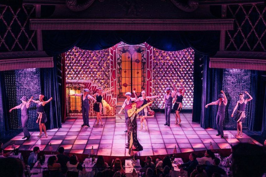 A stage with dancers performing, a patterned backdrop, and an audience seated in front. 2
