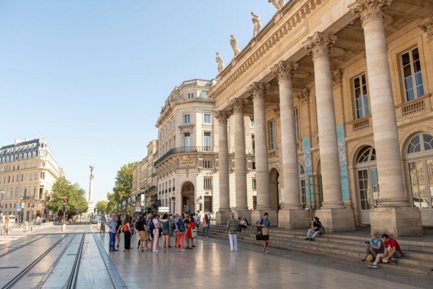A wide boulevard with classical architecture, including columns and statues, and people walking and sitting. 2