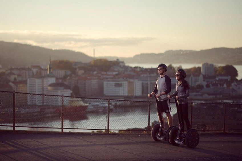 Two people on segways overlooking a cityscape with a body of water and hills in the background. 4