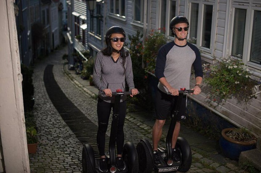 Two people on segways in a narrow street with cobblestone pavement, surrounded by buildings with windows and balconies. 1