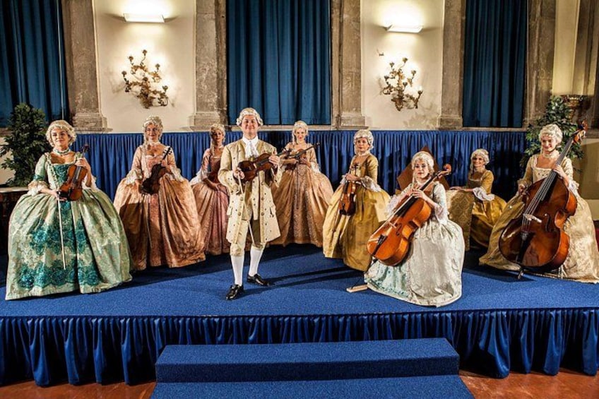 Un groupe de personnes vêtues de costumes d’époque se produit sur une scène dont les rideaux sont bleus, avec une architecture classique en arrière-plan. 2