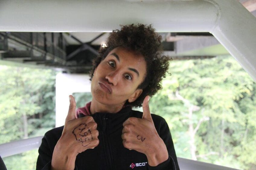 A person with a tattoo on their hand, wearing a black jacket with a logo, standing under a structure with trees in the background. 2