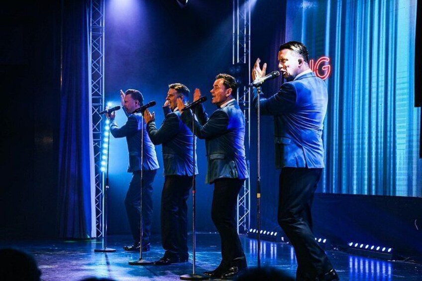 A group of four men singing on a stage with blue lighting. 3